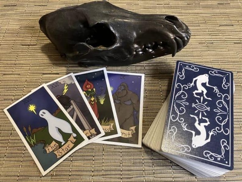 Peut inclure: Un jeu de cartes de tarot avec des illustrations d'un fant&ocirc;me, d'une femme en robe rouge, d'une grande cr&eacute;ature poilue et d'une cr&eacute;ature avec un troisi&egrave;me &oelig;il. Les cartes sont dispos&eacute;es sur un tapis tiss&eacute; avec un cr&acirc;ne d'animal noir en arri&egrave;re-plan.