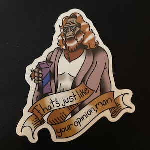 Peut inclure: Un autocollant découpé représentant une figure simiesque brune aux longs cheveux, portant des lunettes de soleil et une robe, tenant un carton violet. Une bannière beige affiche le texte noir : That's just like your opinion, man.
