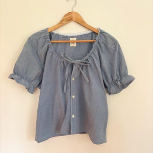 May include: A blue and white gingham blouse with short puff sleeves, a gathered neckline with a tie closure, and a front button placket. The garment features a small white interior label with a circular graphic logo hanging on a wooden hanger.