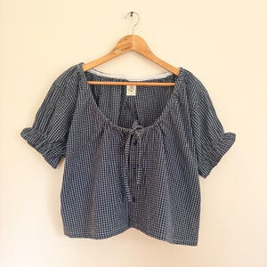 May include: A dark blue and white gingham check blouse featuring short puff sleeves with ruffled cuffs, a wide scoop neckline with a centre tie closure, and a front button placket. A small white label with a line drawing of a flower is visible.