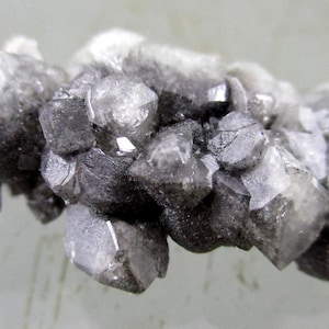 Puede incluir: Un grupo de formaciones minerales cristalinas grises. Los cristales tienen una apariencia facetada, con variaciones en tonos grises claros y oscuros. La estructura general es compacta e irregular, con una textura ligeramente rugosa.