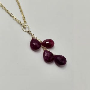May include: A gold-colored chain necklace with a cluster of five deep red, teardrop-shaped gemstones. The gemstones are wire-wrapped and suspended from the chain, creating a delicate and elegant design. The necklace is set against a plain white background.