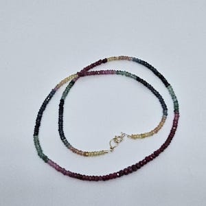 May include: A delicate necklace composed of small, multi-colored beads. The beads transition in color from deep burgundy to shades of pink, green, yellow, and blue, culminating in a dark navy hue. The necklace is secured with a small, silver clasp.