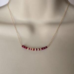 May include: A delicate necklace featuring a strand of faceted, deep red beads interspersed with smaller white and iridescent beads. The beads are strung on a fine, gold-colored chain. The necklace is displayed on a neutral background.