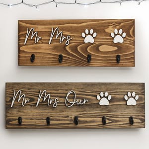 Op de afbeelding: Een houten sleutelhanger met de woorden "Mr. Mrs. Our" en twee witte poot afdrukken. De sleutelhanger heeft vijf haken.