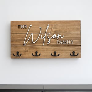May include: A wooden key holder with the text "The Wilson Family" in white lettering. The key holder has four black metal hooks.