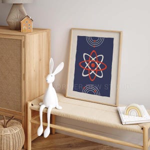 Puede incluir: Una impresi&oacute;n art&iacute;stica enmarcada con un fondo azul marino presenta un dise&ntilde;o de &aacute;tomo blanco y rojo. Una figura de conejo blanco se sienta en un banco tejido junto a la obra de arte enmarcada. Una peque&ntilde;a casa de madera y una l&aacute;mpara est&aacute;n en un mueble de madera.