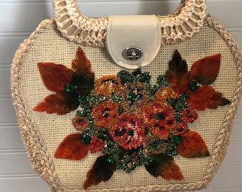 Könnte beinhalten: Eine Strohtasche mit einem geflochtenen Henkel und einem Klappverschluss. Die Vorderseite der Tasche ist mit einem Blumendesign verziert, das orange, rote, grüne und braune Samtblumen und -blätter zeigt. Die Blumen sind mit schillernden Perlen verziert.