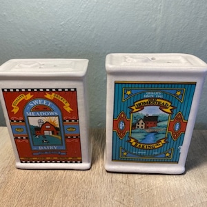 May include: Two white ceramic salt and pepper shakers with a farmhouse theme. The salt shaker says "Sweet Meadows Dairy" and the pepper shaker says "Old Homestead Baking Co."