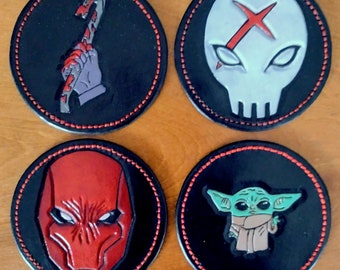 May include: Four round black leather coasters with red stitching. Each coaster features a different graphic design: a red crowbar, a white skull with a red X, a red masked face, and a green alien character.