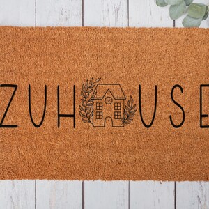 以下が含まれることがあります： 黒い文字で「ZUHAUSE」と書かれた茶色のココナッツ製のドアマット。文字の間に、時計とリースが付いた小さな家が中央に配置されています。