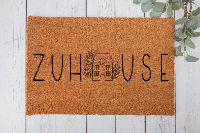 以下が含まれることがあります： 黒い文字で「ZUHAUSE」と書かれた茶色のココナッツ製のドアマット。文字の間に、時計とリースが付いた小さな家が中央に配置されています。