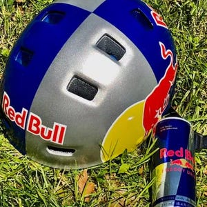 May include: A Red Bull branded helmet in blue, silver, and red, with the Red Bull logo prominently displayed. A Red Bull energy drink can is next to the helmet. The items are on a bed of green grass.