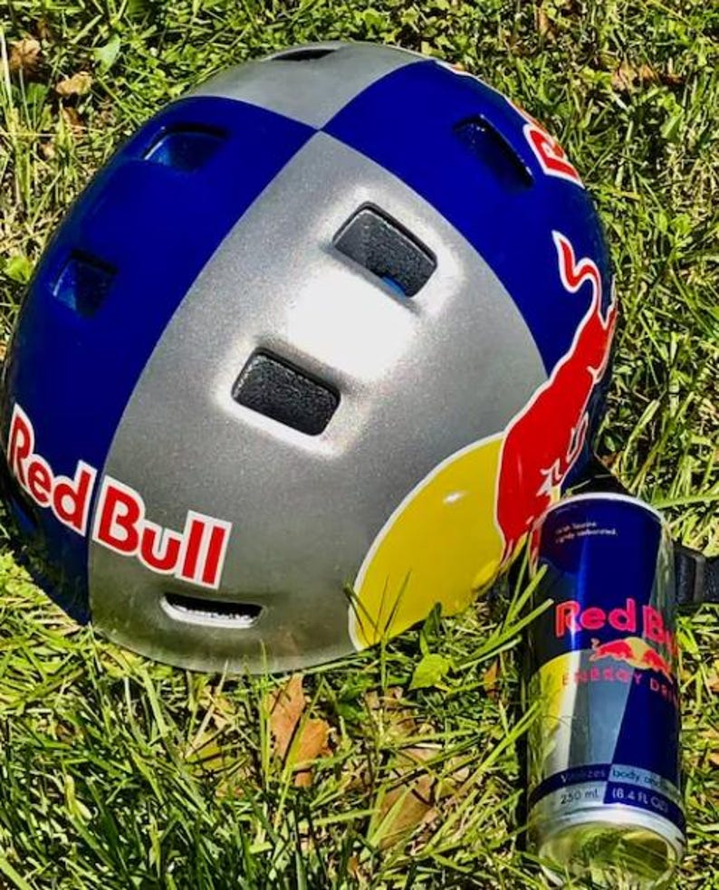 May include: A Red Bull branded helmet in blue, silver, and red, with the Red Bull logo prominently displayed. A Red Bull energy drink can is next to the helmet. The items are on a bed of green grass.
