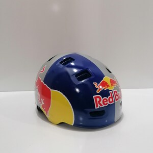 May include: A blue, gray, and yellow Red Bull branded helmet. The helmet has a glossy finish and features the Red Bull logo on the sides. The helmet is designed for sports and outdoor activities.