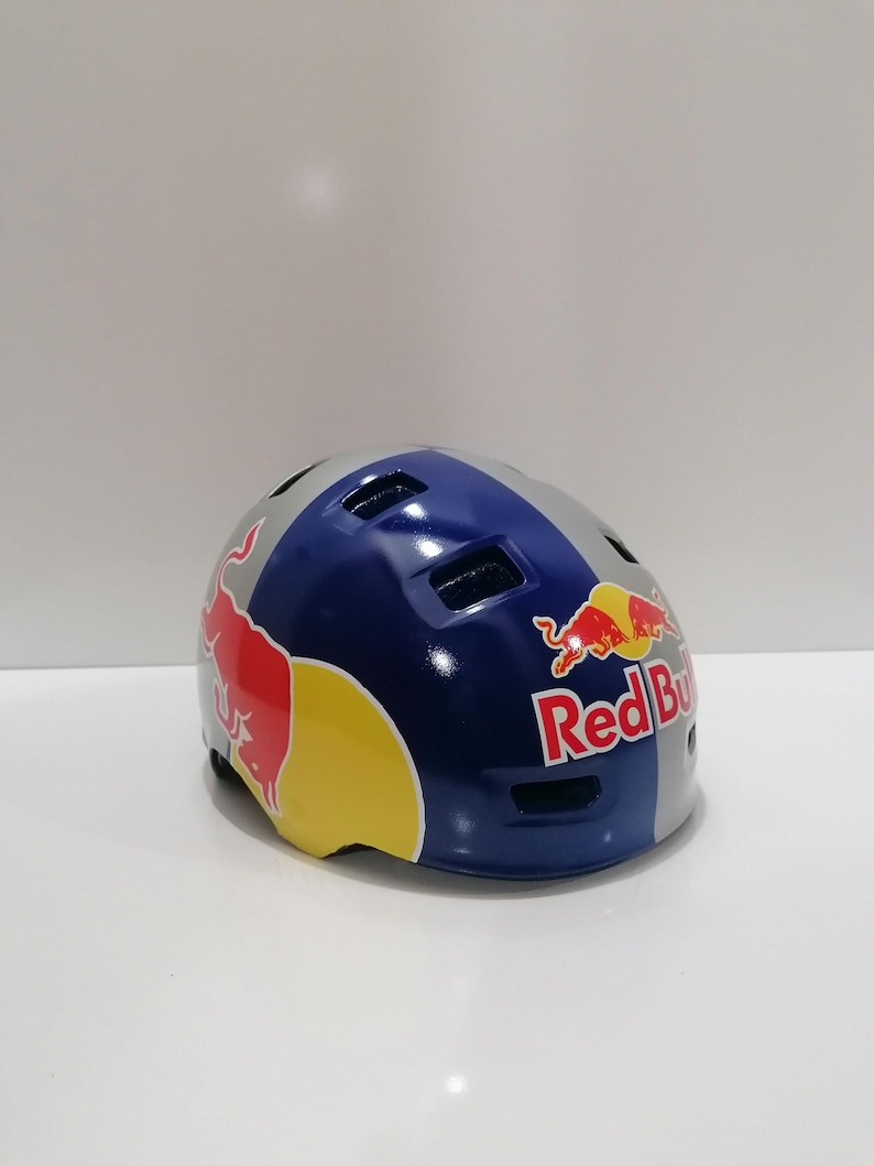May include: A blue, gray, and yellow Red Bull branded helmet. The helmet has a glossy finish and features the Red Bull logo on the sides. The helmet is designed for sports and outdoor activities.