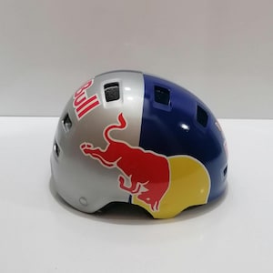 Puede incluir: Un casco Red Bull plateado, azul y amarillo. El casco está dividido en secciones plateadas y azules, con el logotipo de Red Bull en un lugar destacado. El logotipo presenta un toro rojo y la palabra "Bull" en blanco.