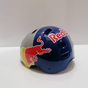 May include: A Red Bull branded helmet. The helmet is primarily blue, gray, and yellow, with the Red Bull logo prominently displayed. The helmet has several ventilation holes and a glossy finish.
