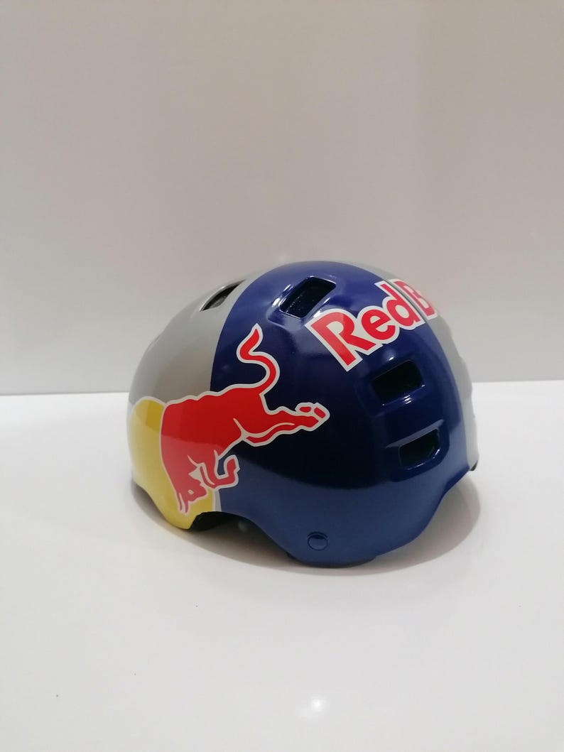 May include: A Red Bull branded helmet. The helmet is primarily blue, gray, and yellow, with the Red Bull logo prominently displayed. The helmet has several ventilation holes and a glossy finish.