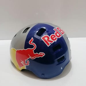May include: A gray, blue, and red Red Bull helmet. The helmet has a glossy finish and features the Red Bull logo on the side and top. The helmet is designed for sports and outdoor activities.