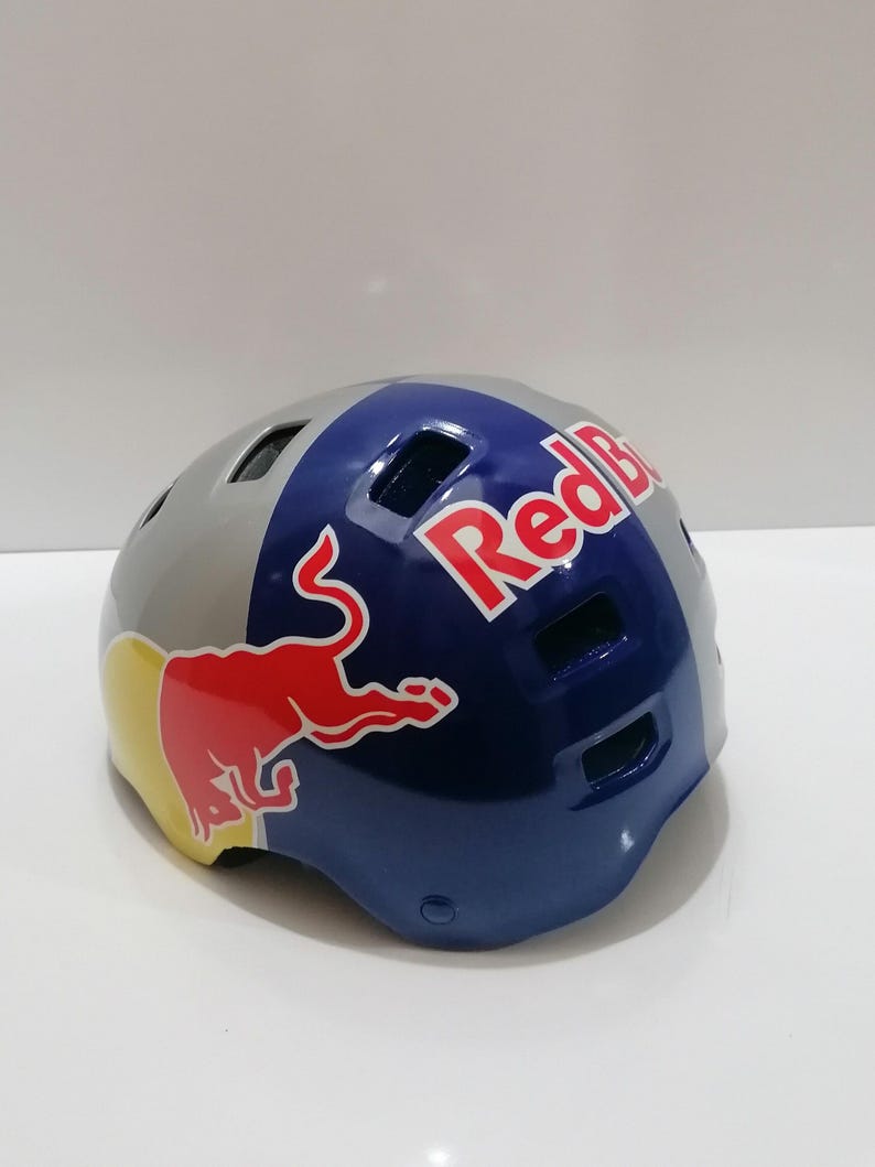 May include: A gray, blue, and red Red Bull helmet. The helmet has a glossy finish and features the Red Bull logo on the side and top. The helmet is designed for sports and outdoor activities.