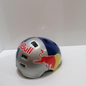 May include: A Red Bull branded helmet with a silver, blue, and yellow color scheme. The helmet features the Red Bull logo in red and white. The helmet is designed for sports and outdoor activities.