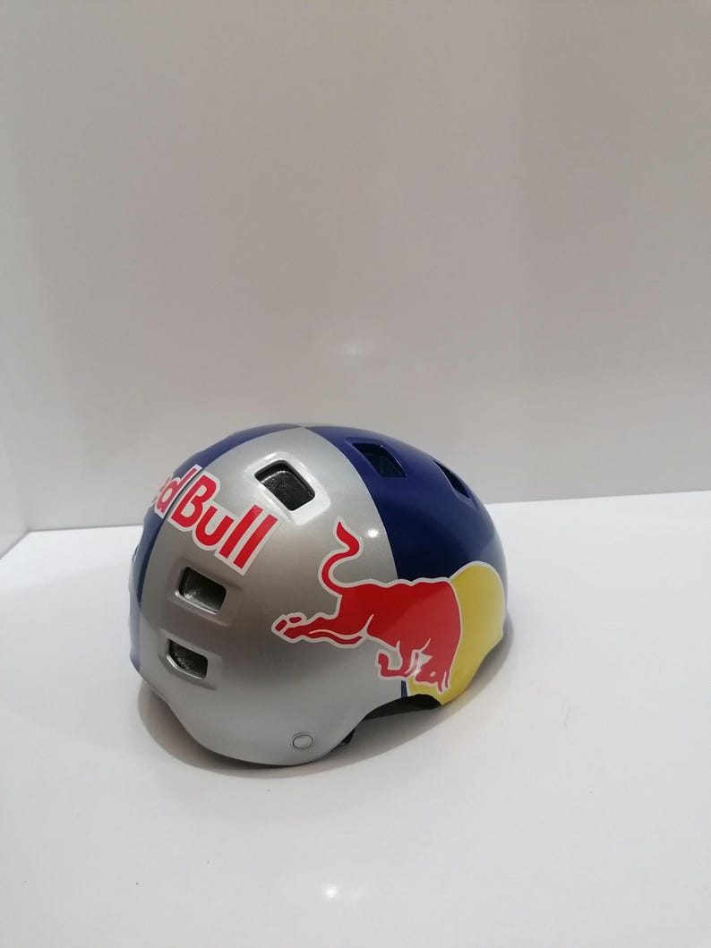May include: A Red Bull branded helmet with a silver, blue, and yellow color scheme. The helmet features the Red Bull logo in red and white. The helmet is designed for sports and outdoor activities.