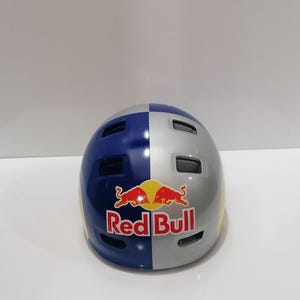May include: A blue, silver, and yellow helmet with the Red Bull logo. The helmet is divided into color-blocked sections and has several ventilation holes. The Red Bull logo is red and yellow with the words "Red Bull" in white.