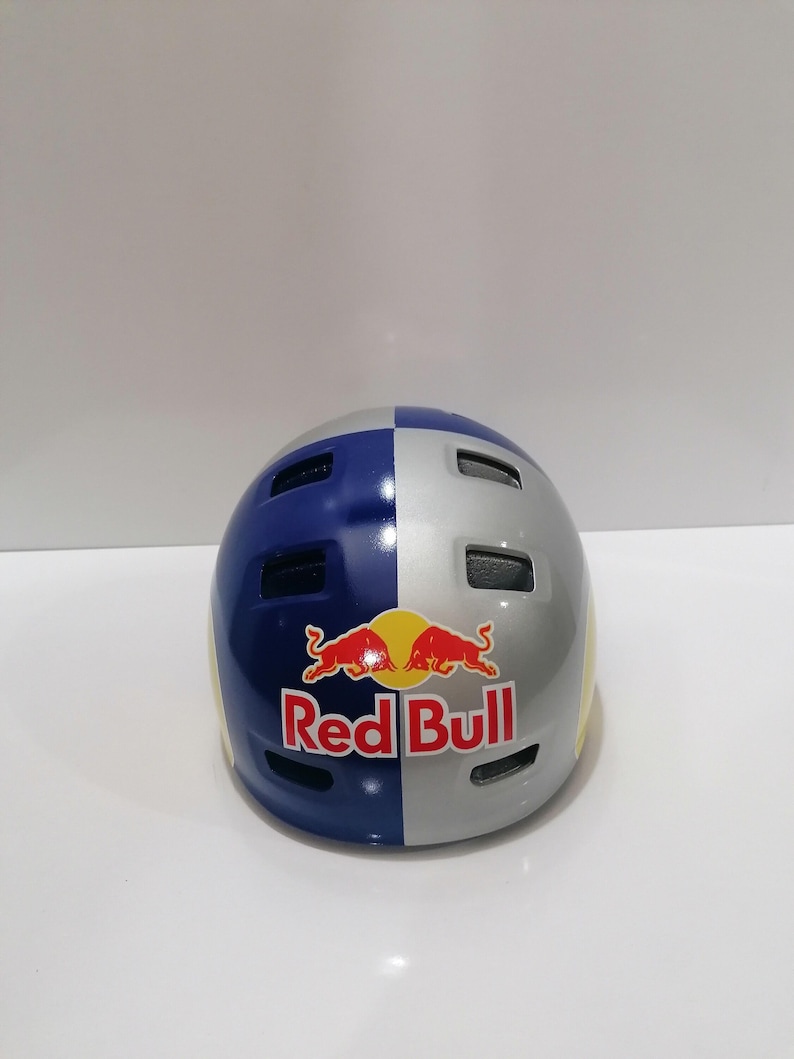 May include: A blue, silver, and yellow helmet with the Red Bull logo. The helmet is divided into color-blocked sections and has several ventilation holes. The Red Bull logo is red and yellow with the words "Red Bull" in white.