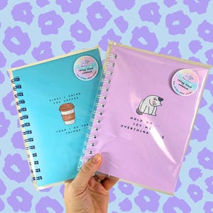 May include: Two spiral bound notebooks with colourful covers. The blue notebook has a coffee cup illustration and the text "First I drink the coffee then I do the things". The pink notebook has a cat illustration and the text "Hold on... Let me overthink this".