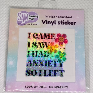 May include: A holographic vinyl sticker with the text "I CAME I SAW I HAD ANXIETY SO I LEFT" and a rainbow glitter background. The sticker has a flower design with a smiley face.
