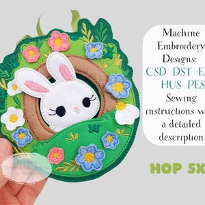 May include: A felt Easter bunny embroidery design with a white bunny head peeking out of a brown circle, surrounded by a green wreath with colorful flowers. The design includes the text "HOP 5X7" and "Machine Embroidery Designs".
