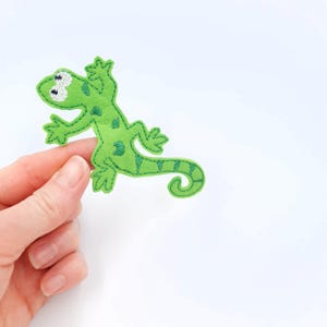 May include: A green felt lizard with embroidered details. The lizard has a light green body with darker green spots and a striped tail. The eyes are white with black details. The lizard is a handmade craft item.