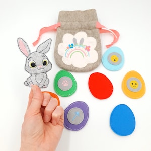 May include: A gray felt Easter egg hunt set with a bunny, a drawstring bag, and colorful felt eggs. The bag features a flower with a rainbow and bunny ears. The eggs are in various colors, some with embroidered details.