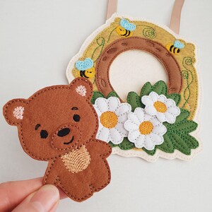 May include: A brown felt teddy bear with embroidered details next to a felt wreath. The wreath features a brown ring, yellow accents, white daisies, green leaves, and blue and yellow bees. A beige ribbon is attached.