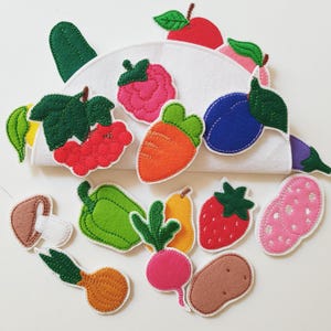 May include: A selection of felt food items in various colours, such as a red apple, orange carrot, and purple plum, displayed around a white felt container. Other items include a green pepper, strawberry, and potato. The items are designed with white stitching.