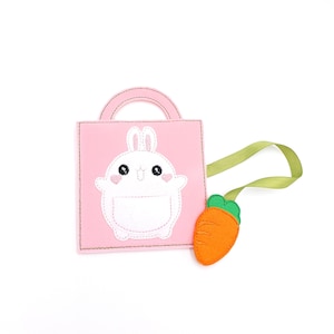 May include: A pink felt bag with a white bunny applique and a green handle. A felt carrot with a green top is attached to the bag with a green ribbon. The bunny has black eyes and a small pocket.