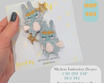 May include: Two light blue bunny hair clips with gold star accents and floral headbands. The clips are on a white card with the text "HOP 4x4" and "FireFly". The card also includes text about machine embroidery designs and sewing instructions.