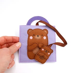 May include: A lavender felt square with a brown bear applique and a smaller bear tag. The bears have smiling faces and pink cheeks. A purple handle and brown ribbon are attached to the square. The item is held by a hand.