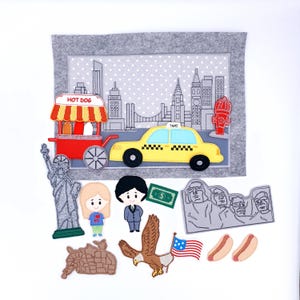 Puede incluir: Un tablero de fieltro con temática de la ciudad de Nueva York, con un taxi amarillo, un puesto de perritos calientes y la Estatua de la Libertad. Los recortes de fieltro adicionales incluyen el Monte Rushmore, un águila y una bandera estadounidense. El fondo es un fieltro gris con un paisaje urbano.