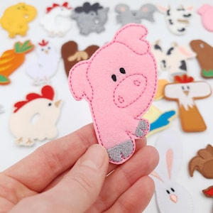 May include: A pink felt pig with grey hooves and black eyes is held in front of a collection of other felt farm animal shapes. Other animals include a chicken, sheep, and cow. The pig is the focal point.