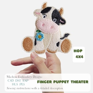 May include: An embroidered finger puppet in the shape of a cow, featuring black and white patches, a pink nose, and a blue flower. The puppet is held up by a hand. Text on the image reads "FINGER PUPPET THEATER" and "HOP 4X4".