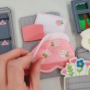 Puede incluir: Escena de casa de muñecas de fieltro con una cama, una manta rosa bordada con rosas blancas. Una mano levanta la manta. Otras piezas de fieltro incluyen un armario, una ventana y un macetero.