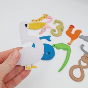 May include: A white felt number five with a yellow beak and feet. Other felt animal numbers and shapes are in the background, including a giraffe, seahorse, whale, and rooster. The items are on a white surface.