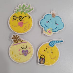 May include: Four whimsical felt appliques. A yellow pineapple with glasses, a blue cloud with a crown, a yellow turnip with a purple heart, and a yellow moon with a nightcap and lantern. Each has a smiling face and is outlined in white.