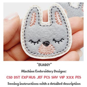 May include: A gray bunny-shaped machine embroidery design with a light pink nose and inner ears, black eyes, and a black stitched outline. The design is labeled "BUNNY" and includes sewing instructions.
