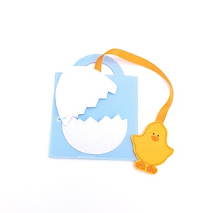 May include: A light blue felt Easter-themed book with a white cracked eggshell design. An orange ribbon is attached to a yellow chick-shaped bookmark. The book has a handle and is on a white background.