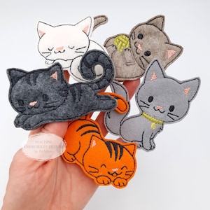 May include: A collection of felt cat-shaped ornaments in various colors, including white, gray, and orange. Each cat has embroidered details, such as stripes, whiskers, and facial expressions. Some cats are curled up, while others are playing with yarn. The text "MACHINE EMBROIDERY DESIGNS by IVAsToys" is visible.