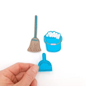 May include: A collection of felt cleaning supplies, including a tan broom with a teal handle, a teal bucket filled with white suds, and a teal dustpan. The dustpan is being held by a hand. All items are on a white background.