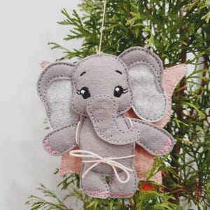 May include: A gray felt elephant ornament with pink accents. The elephant has large gray ears, a trunk, and small pink feet. A pink wing is visible on each side. The ornament is tied with a white string and is hanging from a thin, natural-colored string.